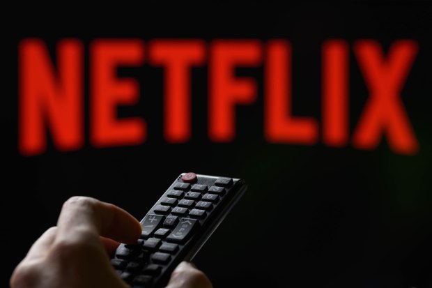 Photo illustration of a hand holding a TV remote with a blurred Netflix logo in the background.
