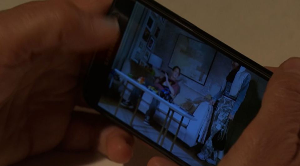 Person holding phone displaying video of two people in a living room.