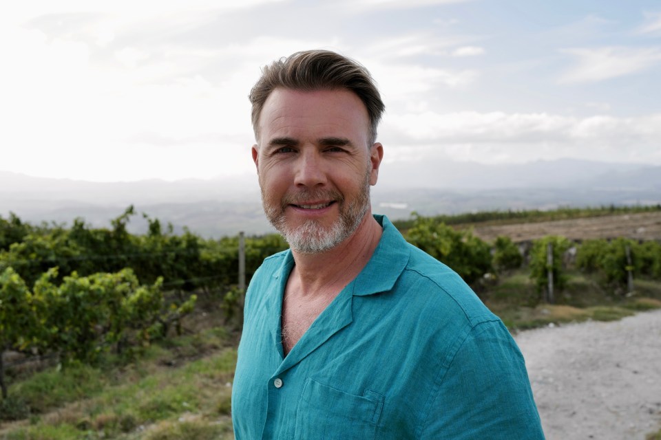 Gary Barlow in a teal shirt, smiling in a vineyard.