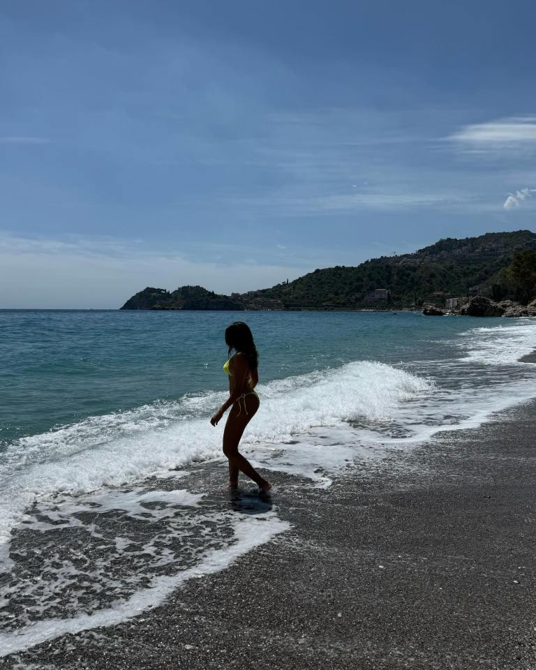 Woman in a yellow bikini wading in ocean waves on a pebble beach.