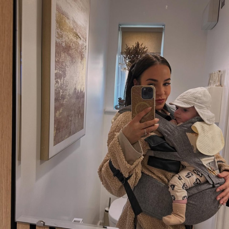 Woman holding baby in carrier takes selfie.
