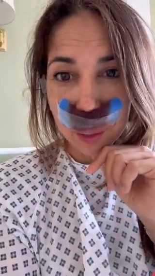 Woman in a hospital gown with a bandage on her nose.