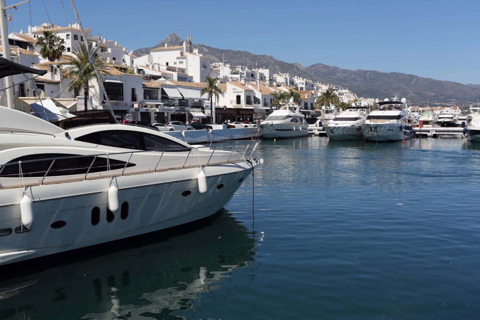 Luxury yachts moored in a marina, with white buildings and mountains in the background.