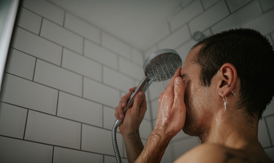 A tattooed man washing his face in a shower.