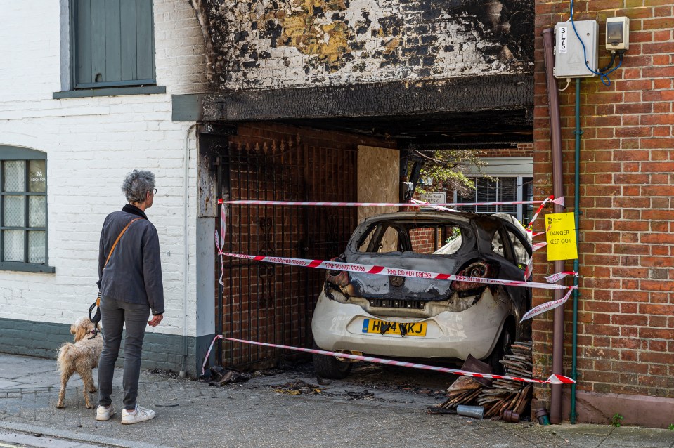 A burnt-out car in a garage, viewed by a woman walking a dog.