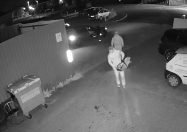 Nighttime security footage of two people walking away from a car, carrying a bag.