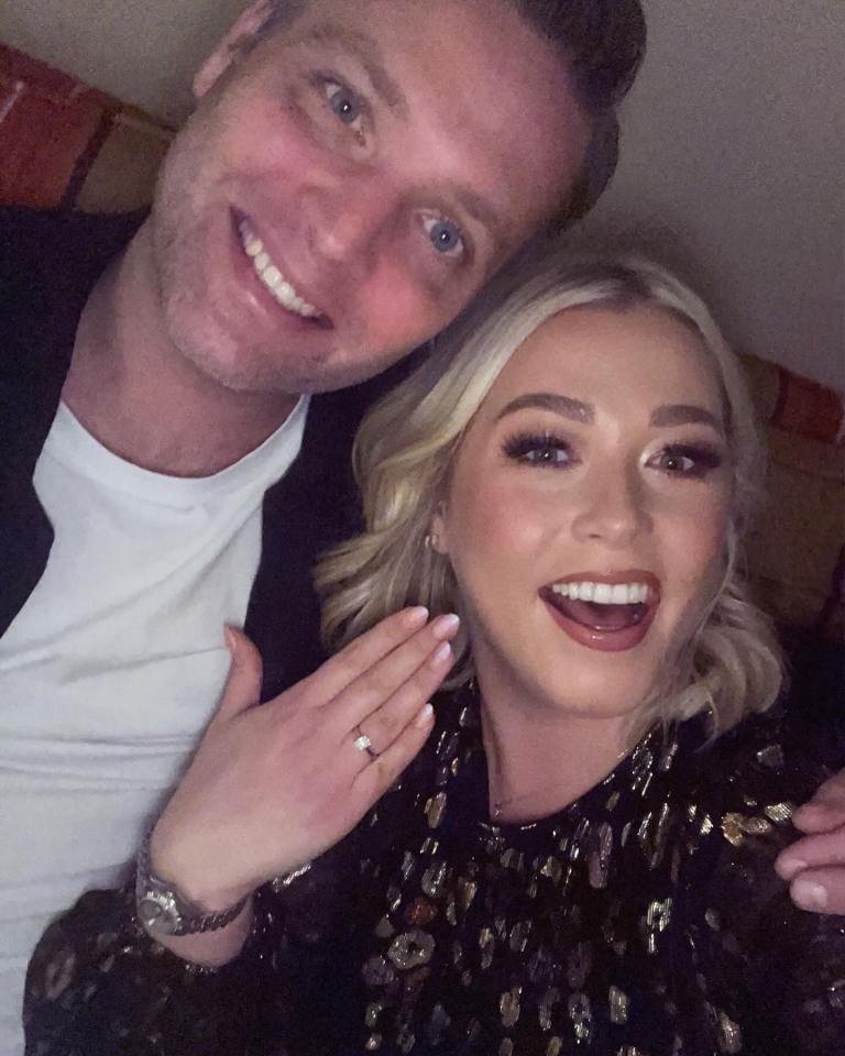 A woman shows off her engagement ring while smiling next to a man.