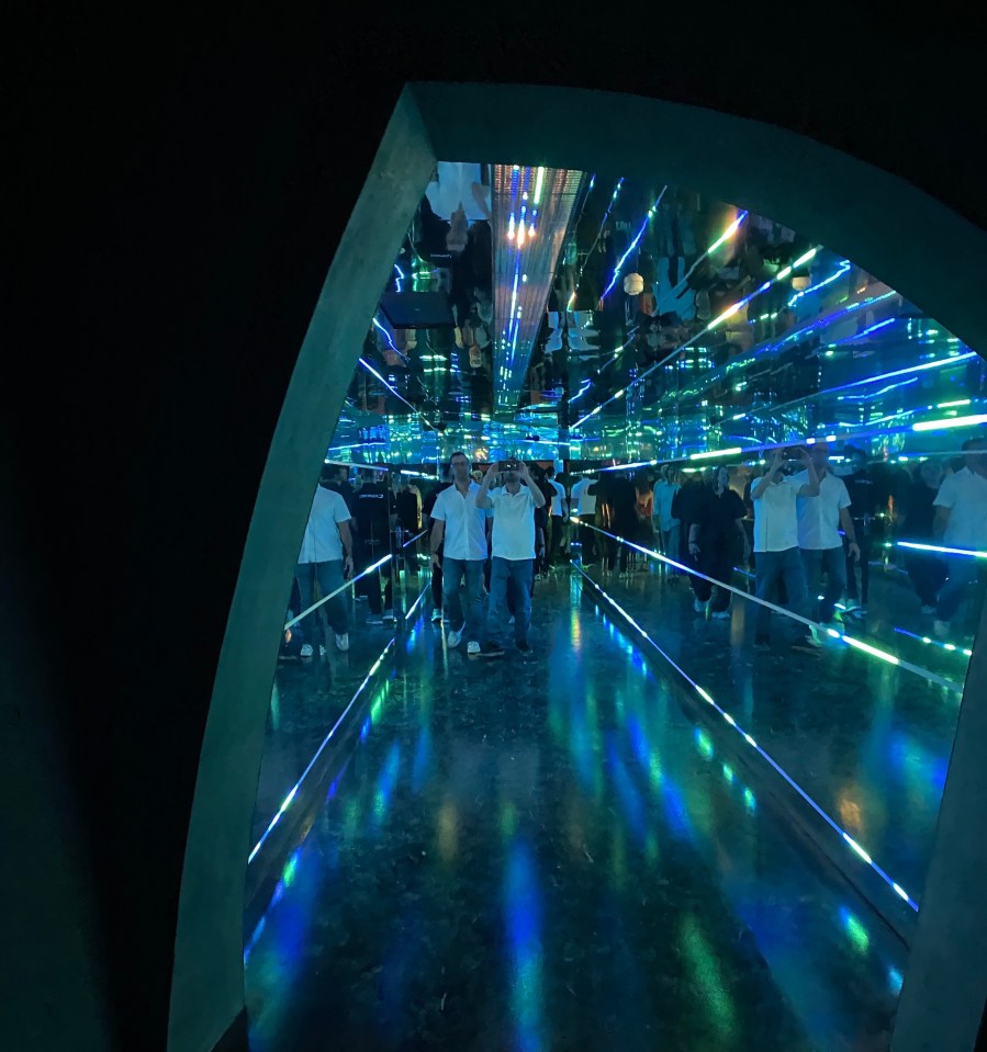 People walking through a mirrored tunnel with blue and green lights.