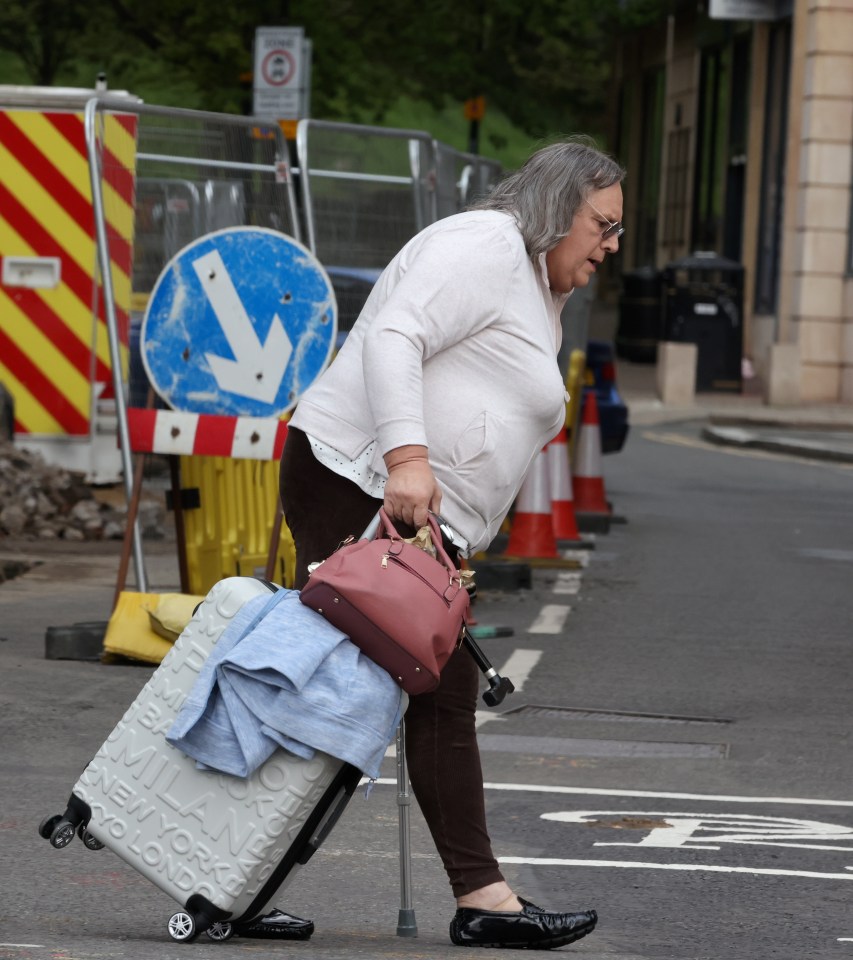 A person with gray hair and glasses uses a cane while walking down a street, pulling a suitcase and carrying a purse.