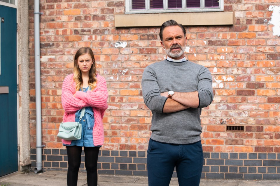 A man and a girl stand against a brick wall, arms crossed.