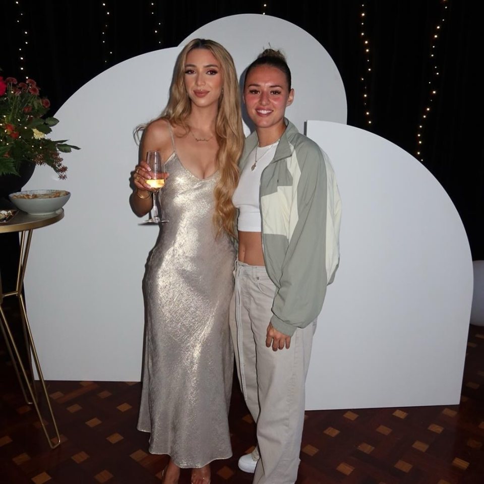 Two women at an event; one in a silver dress, the other in beige pants and a jacket.