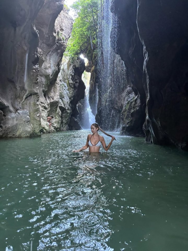 Woman in a bikini wading in a pool of water in a rocky gorge with a waterfall.