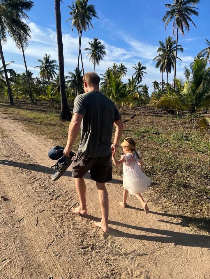 Man and child walking barefoot on a dirt road lined with palm trees.